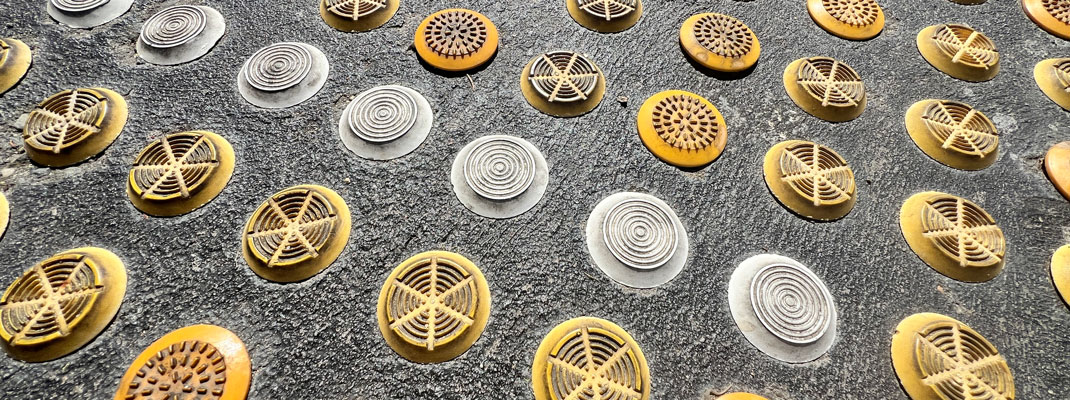 Close-up image of discrete tactile ground surface indicators installed in charcoal concrete, showing multiple circular stud designs and colours including yellow, bronze, and silver. The studs vary in surface pattern and finish, illustrating inconsistent product types, different slip reistabt values and visual contrast within the same warning area.