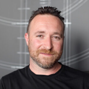 Portrait of Dean Homicki seated against a grey background featuring fine white architectural line drawings. He has short dark hair and a trimmed beard, and wears a black shirt. He looks directly at the camera with a calm, approachable expression.